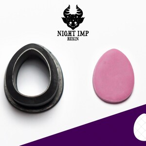 May include: A black silicone mould shaped like an egg, next to a pink egg made of polymer clay. The mould is designed to create a textured egg with a grid pattern.