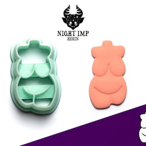 May include: A green plastic cookie cutter shaped like a female body. The cookie cutter is on a white surface. The image also shows a pink cookie in the shape of a female body.