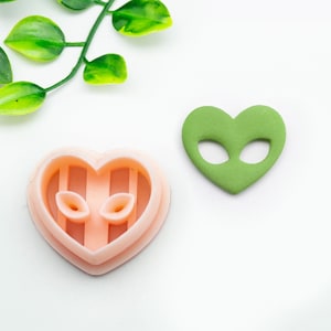 May include: A pink heart-shaped cookie cutter with a design of an alien face inside. Next to it is a green heart-shaped cookie with an alien face cut out.