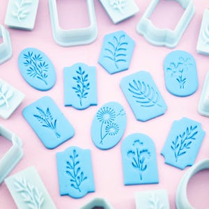 May include: A set of 14 blue plastic cookie cutters featuring various floral and leaf designs. The cutters are arranged on a pink background.