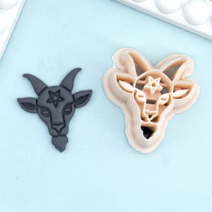 May include: Two goat head cookie cutters. One is a dark gray, solid shape with a pentagram on its forehead. The other is a beige, layered cutter with the same design. Both are on a light blue surface.