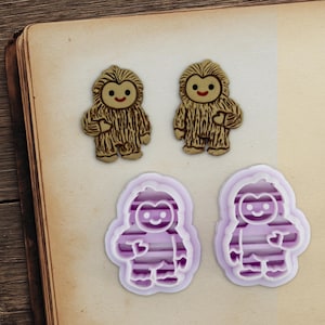 May include: Two Bigfoot-shaped cookie cutters and two Bigfoot-shaped cookies. The cookies are tan with brown fur-like texture and a smiling face. The cookie cutters are purple and white with a striped design.