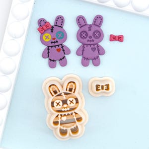 May include: A collection of purple and tan bunny-shaped craft supplies. Two purple bunnies with stitched details and X-shaped eyes and mouths are displayed, along with a small pink bow. A bunny-shaped cookie cutter is also shown.