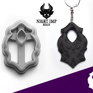 May include: A gray resin mold and a black damask pattern pendant. The mold is in the shape of the pendant, with a cross design. The pendant is attached to a keychain. The Night Imp Resin logo is in the upper left corner.