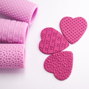 Può includere: Tre tagliabiscotti a forma di cuore rosa con diversi motivi di cuori piccoli. I tagliabiscotti sono su una superficie bianca.