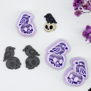 May include: A selection of Halloween-themed cookie cutters. The designs feature a raven perched on a skull, and a raven and skull combination within a purple outline. The raven and skull are in shades of grey and black.