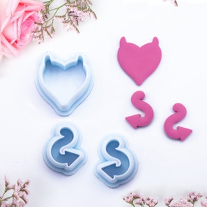 May include: A set of five cookie cutters, including a heart with horns, two swirl shapes, and two heart shapes. The cutters are made of blue plastic and are arranged on a white surface.