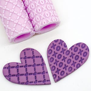 May include: Two polymer clay rollers and two heart-shaped clay pieces. The rollers are light purple with textured patterns of hearts and XOXO. The heart-shaped pieces are purple, one with a grid and heart pattern, the other with an XOXO pattern.