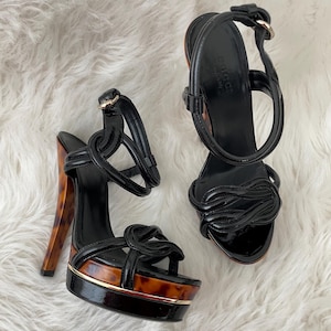 May include: A pair of black patent leather platform sandals featuring a knotted strap design, an adjustable ankle strap with a gold-tone buckle, and a high stiletto heel with a brown and black tortoiseshell pattern and a gold-tone trim.