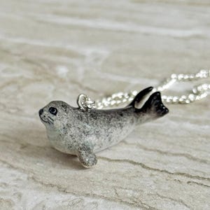 Pu&ograve; includere: Collana con pendente a forma di foca grigia e bianca con catena d'argento. Il ciondolo a forma di foca ha pinne nere e un design maculato. Un affascinante gioiello a tema animale.