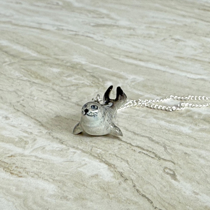 Pu&ograve; includere: Ciondolo in ceramica dipinto a mano a forma di foca, con catenina d'argento. La foca &egrave; grigia con pinne e orecchie nere e occhi blu. Un accessorio stravagante a tema animale.