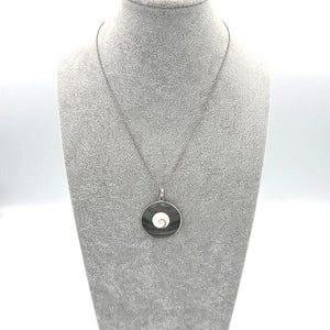 May include: A silver necklace with a circular pendant. The pendant features a dark gray outer ring and a white spiral design in the center. The necklace is displayed on a gray mannequin.