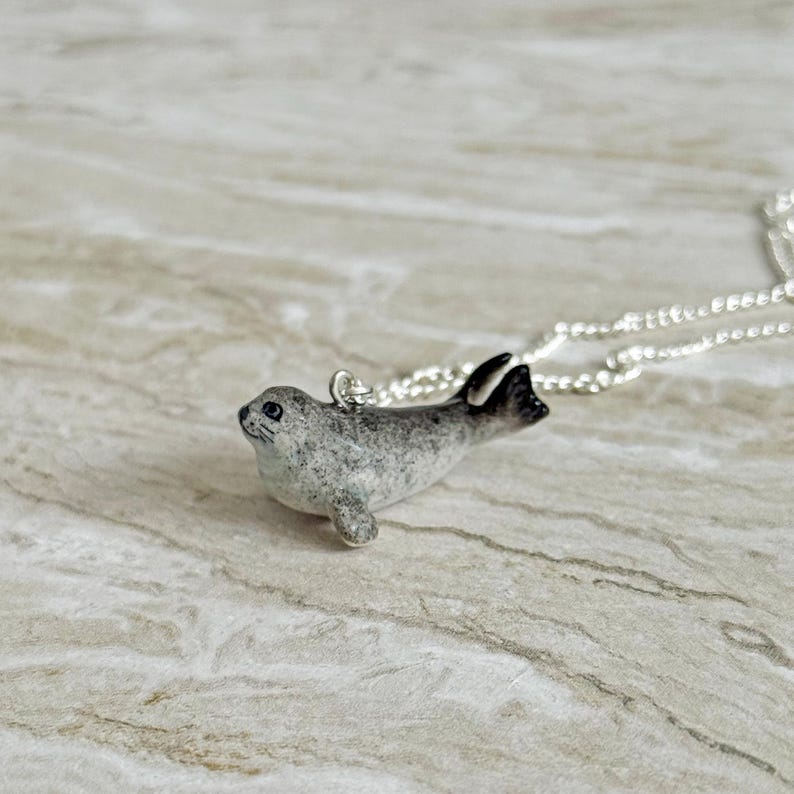 Pu&ograve; includere: Collana con pendente a forma di foca grigia e bianca con catena d'argento. Il ciondolo a forma di foca ha pinne nere e un design maculato. Un affascinante gioiello a tema animale.