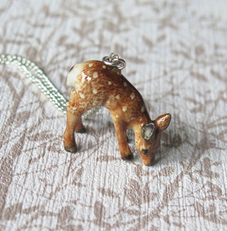 May include: A brown and white ceramic deer pendant on a silver chain. The deer is looking down and has a spotted pattern.