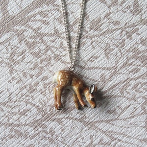 May include: A silver chain necklace with a brown and white ceramic deer pendant. The deer is looking down and has a spotted pattern.