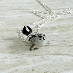 Pu&ograve; includere: Una collana d'argento con un piccolo ciondolo a forma di foca. La foca &egrave; grigia e bianca con pinne nere e un volto sorridente. La catena &egrave; delicata e argentata, creando un affascinante accessorio a tema marino.
