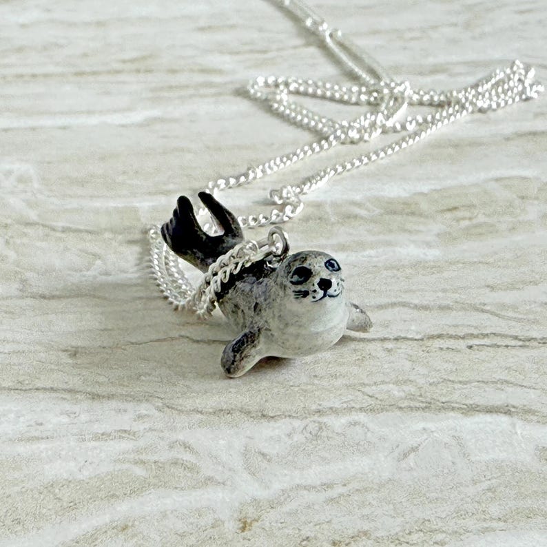 Pu&ograve; includere: Una collana d'argento con un piccolo ciondolo a forma di foca. La foca &egrave; grigia e bianca con pinne nere e un volto sorridente. La catena &egrave; delicata e argentata, creando un affascinante accessorio a tema marino.