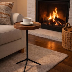 May include: A small, round side table with a dark wood top and a black metal base. A white teacup and saucer sit on the table. The table is next to a sofa and a fireplace with a burning fire.