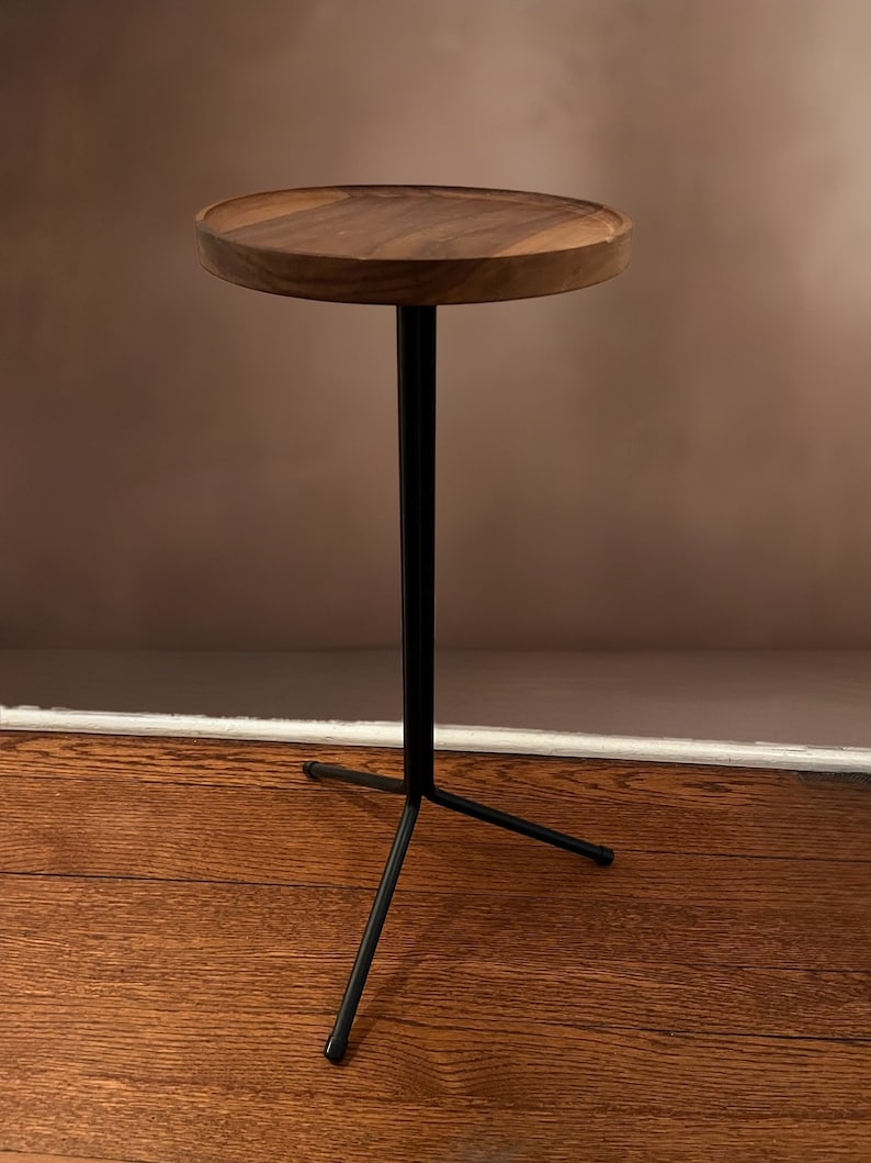 May include: A small, round side table with a dark wood top and a black metal base. The table has a single central support and three legs, creating a tripod-like structure. The wooden tabletop is approximately 30 cm in diameter.