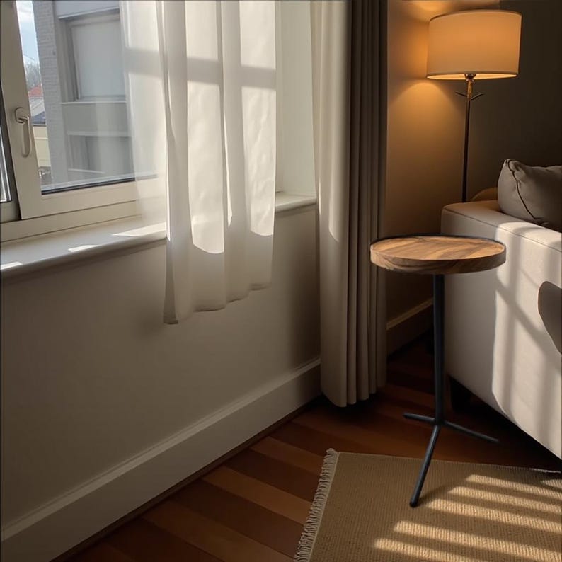 May include: A small, round wooden side table with a dark metal tripod base. The table is positioned next to a white sofa and a window with sheer white curtains. The room has hardwood floors and a beige rug.