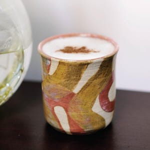 May include: A ceramic mug filled with a creamy beverage, topped with a sprinkle of cinnamon. The mug features a unique design with a mix of colors, including brown, yellow, and red. The rim of the mug is a soft pink.