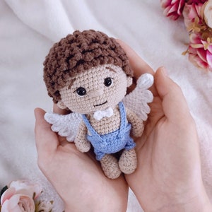 May include: A crocheted angel doll with brown hair, white wings, and a blue outfit. The doll is being held in the hands of a person.