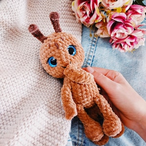 May include: A brown crocheted plush toy with large blue eyes and long antennae. The toy is sitting on a denim and knitted fabric background.