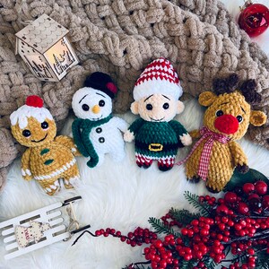 May include: Four crocheted Christmas ornaments: a gingerbread person, a snowman, an elf, and a reindeer. The ornaments are on a white fluffy surface with red berries and a wooden sign that says "Xmas".