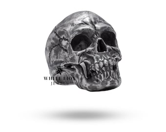 May include: A detailed silver skull ring with a textured surface. The skull has a pronounced jawline and teeth. The ring is a statement piece for those who appreciate gothic and macabre jewelry.