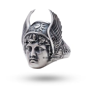 May include: A silver ring with a detailed design of a winged helmet with a sun face on the front. The ring is engraved with the words "White Lion Jewels".
