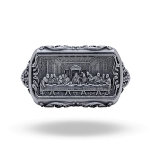 Può includere: Un anello rettangolare color argento con un dettagliato bassorilievo dell'Ultima Cena sulla parte frontale. La superficie metallica presenta una finitura ossidata con intricati motivi a volute lungo i bordi e i lati dell'anello.