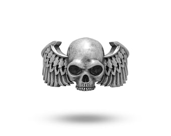 May include: A silver-toned ring featuring a central human skull design flanked by detailed feathered wings on both sides. The metal has a weathered, oxidized finish that highlights the deep contours of the skull and the individual feathers.