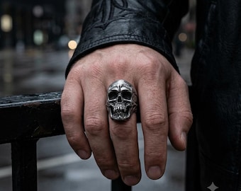 May include: A silver-toned ring featuring a detailed, realistic human skull design. The skull has deep-set eye sockets, a defined nasal cavity, and visible teeth. The metal surface has a polished finish with darkened recessed areas to highlight the intricate anatomical features of the skull structure.