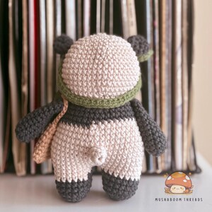 Puede incluir: Mu&ntilde;eca de oso panda de ganchillo hecha a mano con cabeza beige, brazos y piernas grises y cuerpo blanco y gris. El panda lleva una bufanda verde y una bandolera beige. La mu&ntilde;eca se exhibe frente a una pila de discos de vinilo.