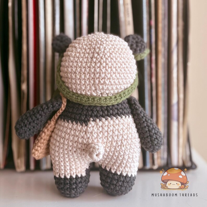 Puede incluir: Mu&ntilde;eca de oso panda de ganchillo hecha a mano con cabeza beige, brazos y piernas grises y cuerpo blanco y gris. El panda lleva una bufanda verde y una bandolera beige. La mu&ntilde;eca se exhibe frente a una pila de discos de vinilo.