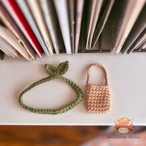 Puede incluir: Una diadema de ganchillo hecha a mano con detalles de hojas en verde oliva y una peque&ntilde;a bolsa de ganchillo marr&oacute;n con asa. Los art&iacute;culos se muestran sobre una superficie blanca con un fondo borroso de libros.