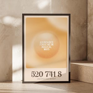 May include: A framed poster in a warm, neutral colour scheme. The poster displays the text "Abundance Finds Me in Magical Ways" and the numbers "520 741 8" with "Unexpected Money" below. The background has a soft, blurred effect.