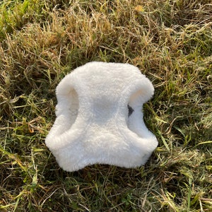 May include: White faux fur dog vest with two straps. The vest is lying on a bed of green grass.