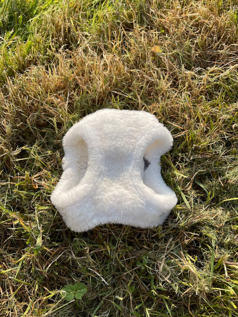 May include: White faux fur dog vest with two straps. The vest is lying on a bed of green grass.