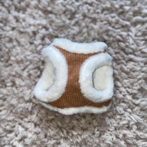 May include: A brown and white dog harness with a soft, fuzzy lining. The harness is made of a knitted fabric and has a white faux fur trim.