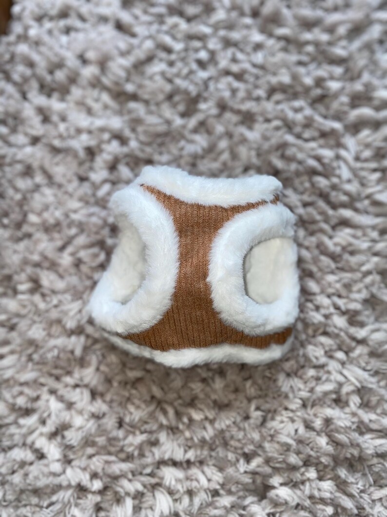 May include: A brown and white dog harness with a soft, fuzzy lining. The harness is made of a knitted fabric and has a white faux fur trim.