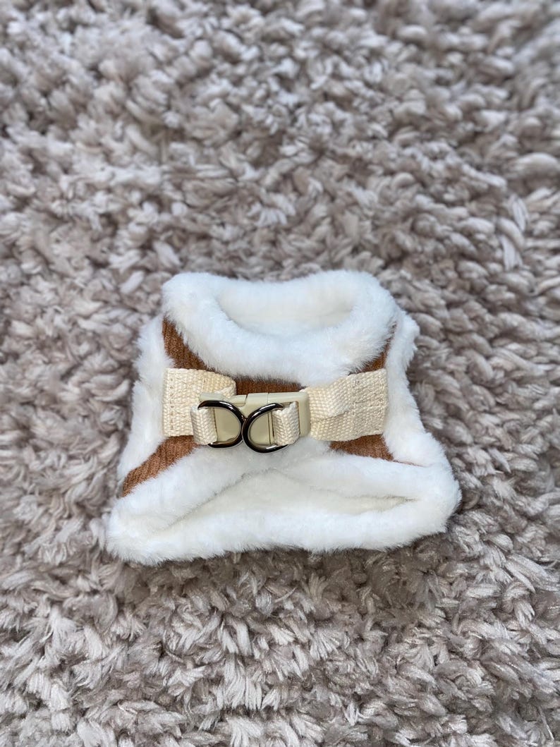 May include: A brown and white faux fur dog harness with a white buckle and two metal rings.