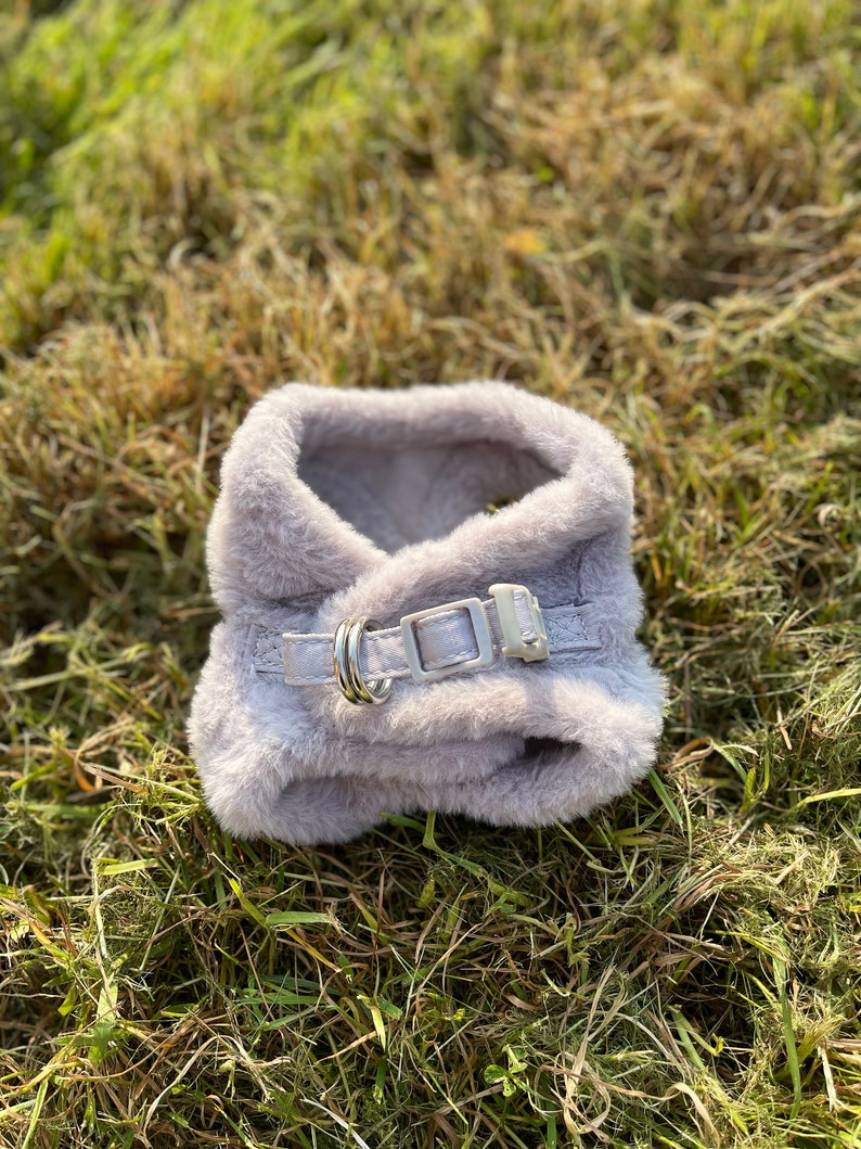 May include: A soft, gray, faux fur dog harness with a silver buckle and two silver rings.