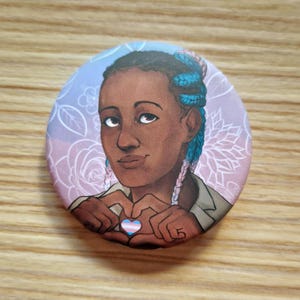 Op de afbeelding: Ronde button met een illustratie van een persoon met blauwe vlechten die met hun handen een hartvorm vormen. Het hart is gekleurd met de transgender pride vlag. De achtergrond is lichtpaars met bloemmotieven.