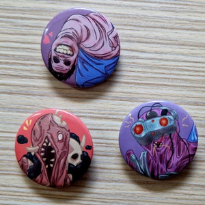 Peut inclure: Trois badges ronds avec différents personnages de dessins animés. Le premier badge représente un personnage de dessin animé rose et bleu avec un large sourire. Le deuxième badge représente un personnage de dessin animé rouge et noir avec une grande bouche ouverte. Le troisième badge représente un personnage de dessin animé violet avec une grosse tête et deux yeux rouges.