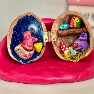 Puede incluir: Diorama caprichoso de cáscara de nuez con bisagras. Un lado presenta un ratón rosa en una taza floral, el otro un pájaro morado. También hay libros en miniatura y una seta. El diorama está sobre una base rosa.