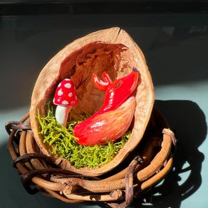 Peut inclure: Une scène miniature fantaisiste avec un cerf rouge et un champignon à pois blancs, nichés dans une coquille de noix. La coquille repose dans un panier tressé, avec de la mousse verte.