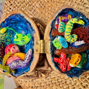 Pode incluir: Um diorama artesanal de casca de noz aberta com uma dobradiça de latão. O interior é pintado com uma cena oceânica azul, apresentando criaturas marinhas e corais esculpidos coloridos. O diorama está sobre um fundo tecido.