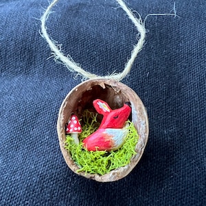 May include: A small, red wooden fox figurine with closed eyes is nestled in a halved walnut shell. The shell is filled with green moss and a small red and white mushroom. A twine loop is attached to the top of the shell for hanging.