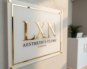 May include: A white rectangular sign with gold trim and lettering. The sign reads "LXN Aesthetics Clinic London" in gold. The sign is mounted on a light-coloured wall with gold fixings.